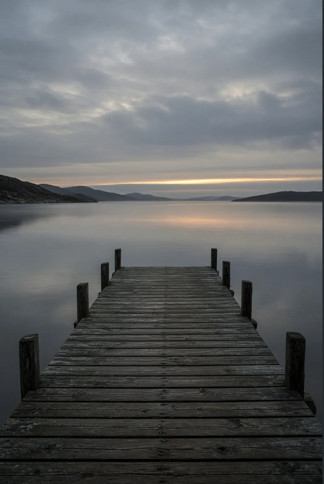 A dark wooden pier reaching into still water at first light
